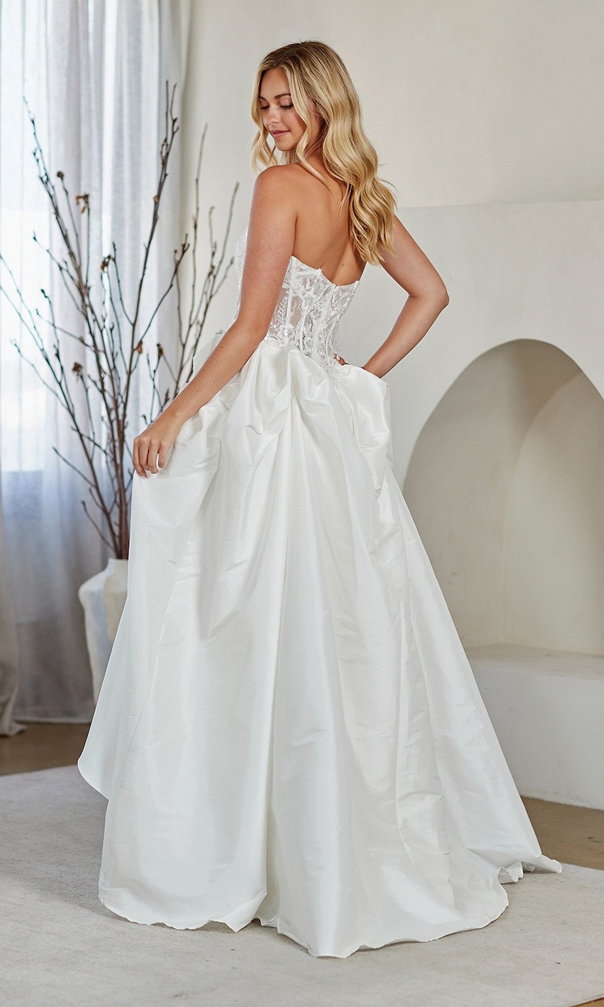 A woman in the Amelia Couture 2087 long prom dress with a strapless lace corset bodice and full A-line skirt stands indoors, glancing over her shoulder.